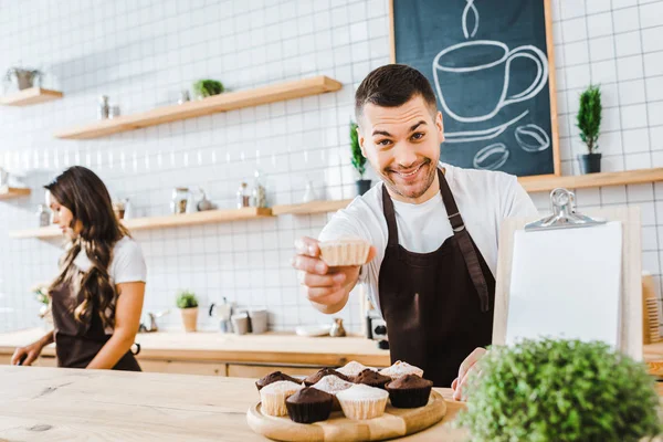 Enfoque selectivo de barista guapo dando cupcake wile atractiva cajera morena trabajando en la cafetería - foto de stock