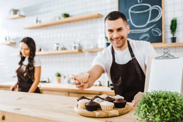 Enfoque selectivo de barista guapo dando cupcake cajero astuto trabajando en la cafetería - foto de stock