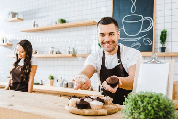 Messa a fuoco selettiva del cassiere bello in grembiule dando cupcakes wile barista lavorando in caffetteria — Foto stock