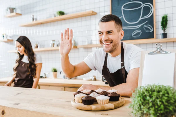 Selektiver Fokus der hübschen Kassiererin, die hinter der Theke steht und lächelt, während ein attraktiver Barista im Kaffeehaus arbeitet — Stockfoto