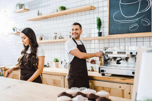 Barista haciendo café y sonriendo astuto cajero atractivo trabajando detrás del mostrador de la barra en la cafetería - foto de stock