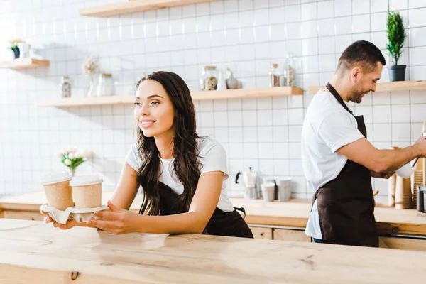 Attractive brunette cashier standing behind bar counter and holding paper cups wile barista working in coffee house — Stock Photo