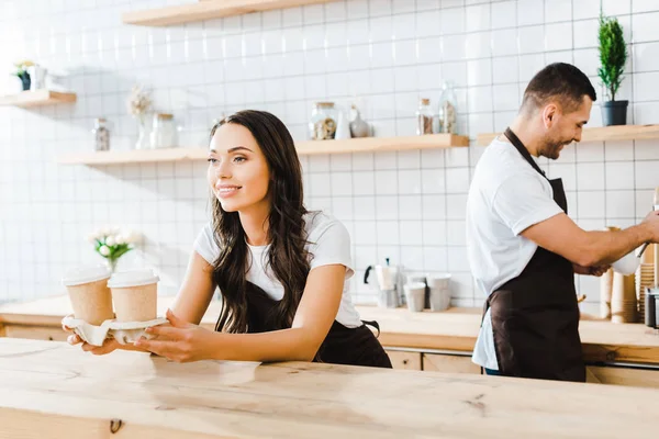 Atractiva cajera morena de pie detrás del mostrador de la barra y sosteniendo vasos de papel barista wile trabajando en la cafetería - foto de stock