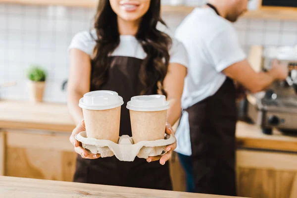 Corte vista de morena caixa de pé e segurando copos de papel wile barista trabalhando em casa de café — Fotografia de Stock