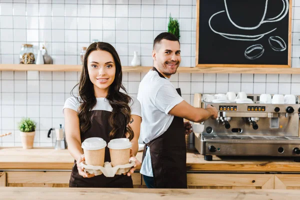 Brunette cashier standing and holding paper cups wile man making coffee in coffee house — Stock Photo