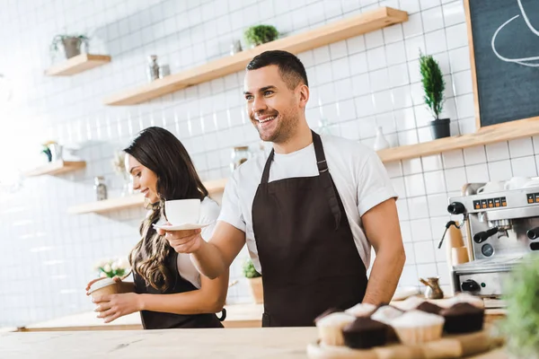 Handsome cashier holding cup and saucer wile attractive brunette barista closing paper cup in coffee house — Stock Photo