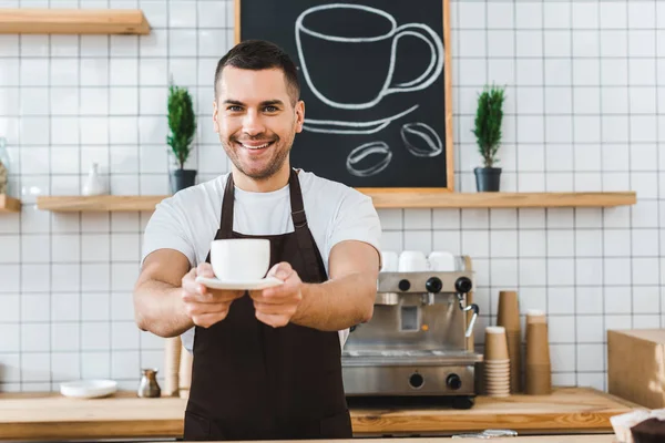 Handsome barista in brown apron holding cup and saucer in coffee house — Stock Photo