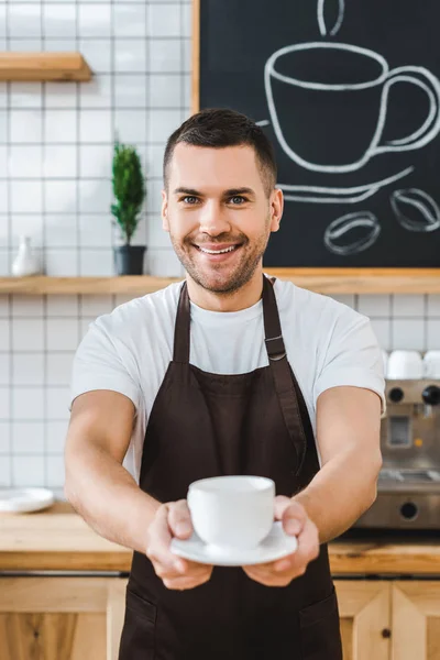 Messa a fuoco selettiva di bel barista in grembiule marrone che tiene tazza e piattino in caffetteria — Foto stock