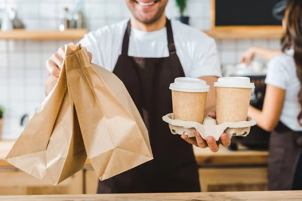 Cropped view of cashier holding paper cups and bags wile brunette barista making coffee in coffee house — Stock Photo
