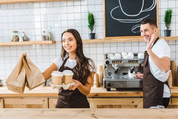 Atractiva morena cajera sosteniendo vasos de papel y bolsas wile barista ondeando en la cafetería - foto de stock