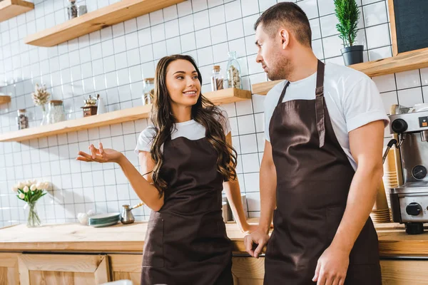 Cajeros en delantales hablando cerca de mostrador de bar de madera en la cafetería - foto de stock