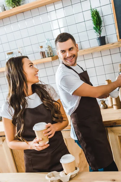 Barista in brown apron making coffee and attractive brunette cashier holding paper cup in coffee house — Stock Photo