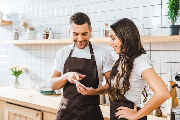 Cashiers in aprons standing near bar counter and looking to smartphone in coffee house — Stock Photo