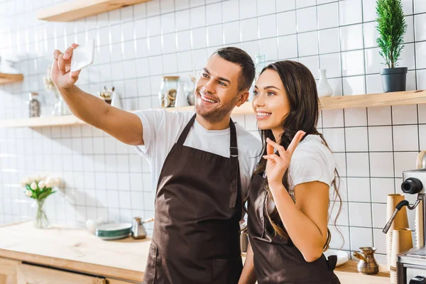 Cashiers in aprons standing near bar counter and taking selfie in coffee house — Stock Photo