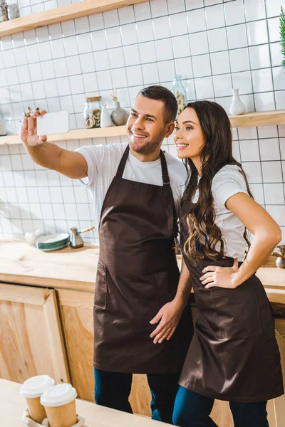 Cajeros en delantales de pie y tomando selfie en la cafetería - foto de stock