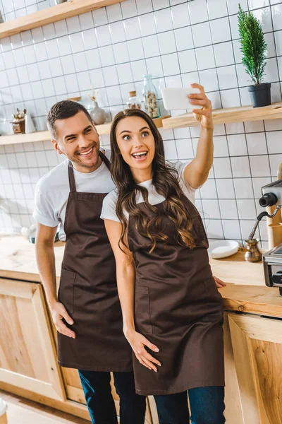 Cassieri in grembiuli in piedi, sorridenti e scattare selfie in caffetteria — Foto stock