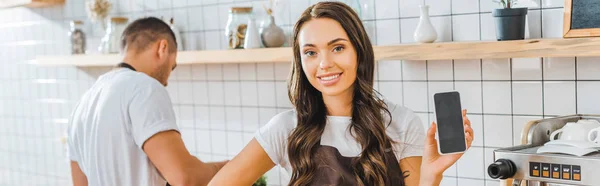 Atractivo cajero mostrando teléfono inteligente con pantalla en blanco barista wile trabajando en la cafetería - foto de stock