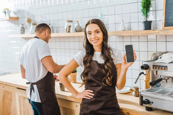 Attractive brunette cashier showing smartphone with blank screen wile barista working in coffee house — Stock Photo