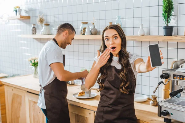 Überraschte brünette Kassiererin zeigt Smartphone mit leerem Bildschirm wile Barista arbeitet im Kaffeehaus — Stockfoto