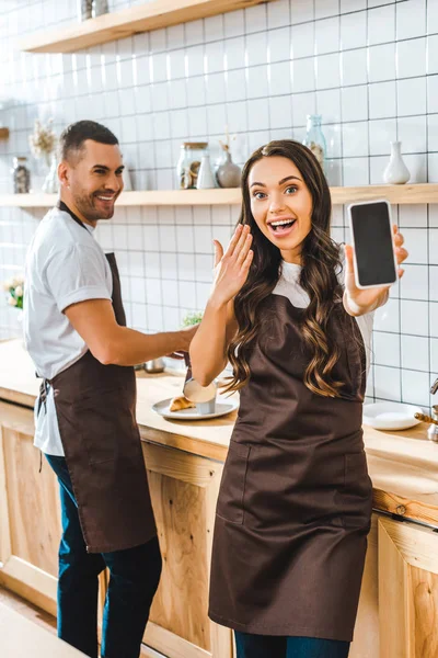 Caixa morena mostrando smartphone com tela em branco wile barista sorrindo na casa de café — Fotografia de Stock