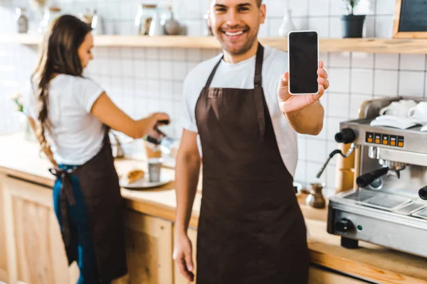 Messa a fuoco selettiva del cassiere bello mostrando smartphone con schermo bianco wile barista che lavora in caffetteria — Foto stock