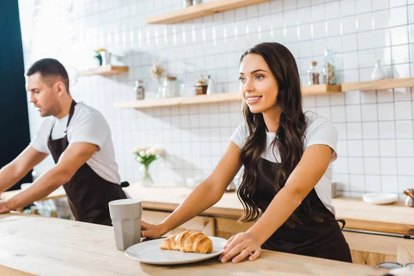 Caixa atraente de pé atrás do balcão de bar com copo, prato e croissant wile barista trabalhando na casa de café — Stock Photo