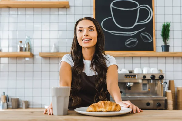 Attractive cashier standing behind bar counter with cup, plate and croissant in coffee house — Stock Photo