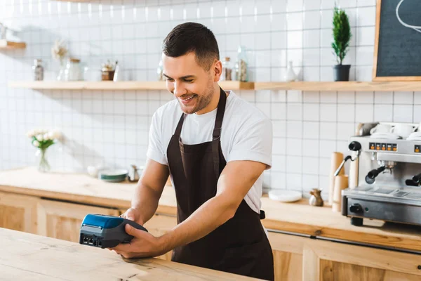 Schöne Kassiererin steht in brauner Schürze neben Theke und hält blaues Terminal im Kaffeehaus — Stockfoto