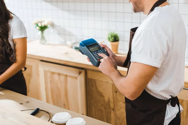 Cropped view of cashier holding blue terminal wile brunette barista working in coffee house — Stock Photo