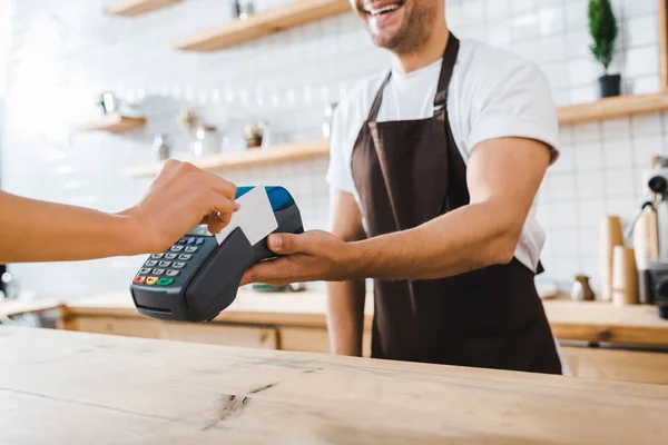 Vista recortada de cajero de pie cerca del mostrador de bar y la celebración de la mujer terminal astuta que paga con tarjeta de crédito en la cafetería - foto de stock