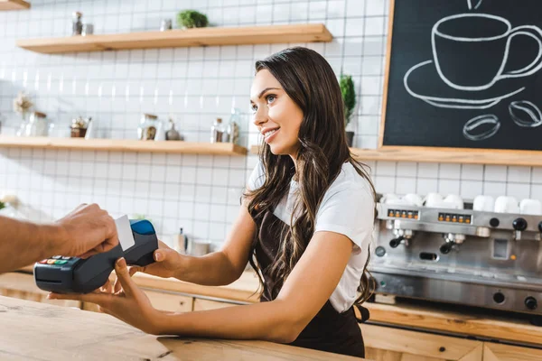 Attraente cassiere in piedi vicino bancone bar in grembiule marrone e tenendo terminale uomo astuto che paga con carta di credito in caffetteria — Foto stock