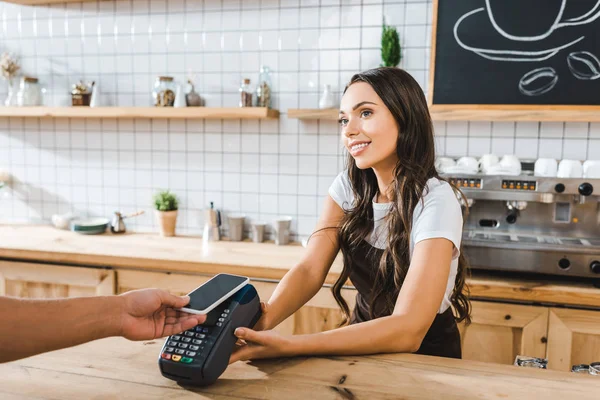 Attraente cassiere in piedi vicino bancone bar in grembiule marrone e tenendo terminale uomo astuto pagare con smartphone in caffetteria — Foto stock