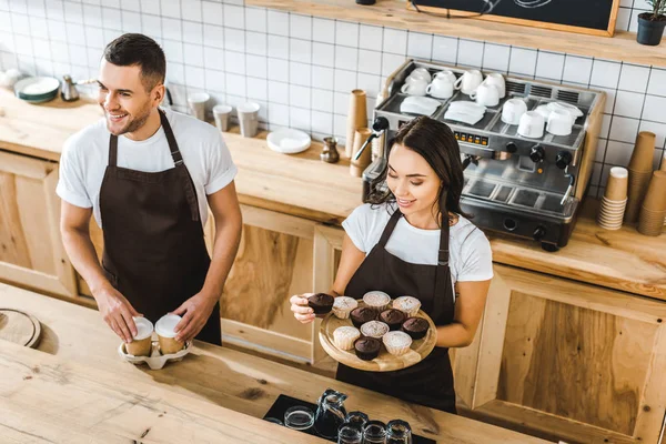 Attractive cashier with cupcakes and handsome barista with paper cups standing behind bar counter in coffee house — Stock Photo