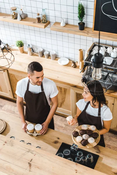 Atractiva cajera con cupcakes y barista guapo con copas de papel de pie en delantales marrones detrás de barra mostrador en la cafetería - foto de stock