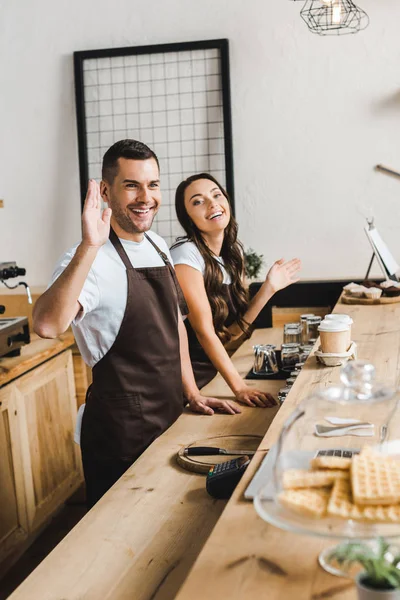 Cashiers smiling and waving behind wooden bar counter in coffee house — Stock Photo