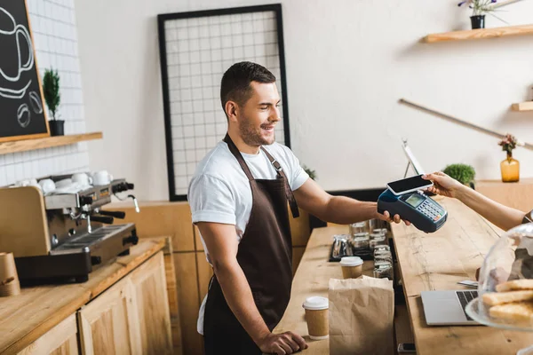 Handsome barista standing near bar counter in brown apron and holding terminal wile woman paying with smartphone in coffee house — Stock Photo