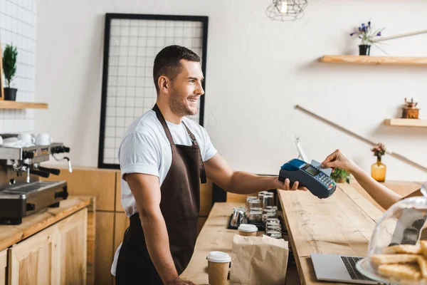 Handsome cashier standing in brown apron and holding terminal wile woman paying with credit card in coffee house — Stock Photo