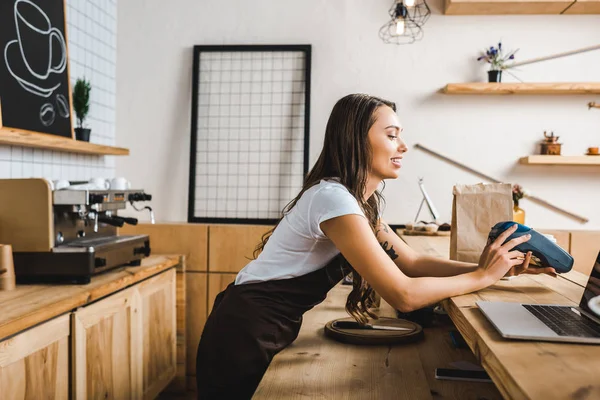 Attractive cashier standing in brown apron and holding terminal behind bar counter in coffee house — Stock Photo