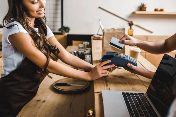 Vista cortada de caixa de pé em avental marrom e segurando homem wile terminal pagando com cartão de crédito na casa de café — Fotografia de Stock