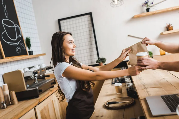 Attractive cashier in brown apron giving paper cup and bag to man in coffee house — Stock Photo