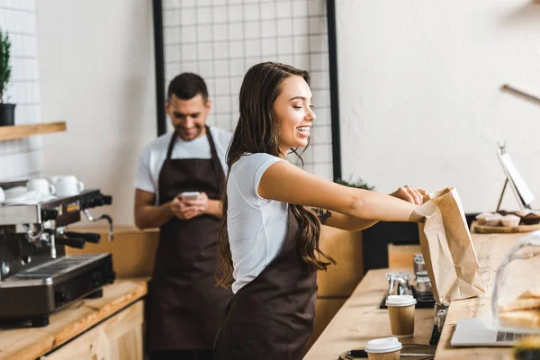 Attractive cashier in brown apron packing paper bag wile barista looking to smartphone near bar counter in coffee house — Stock Photo