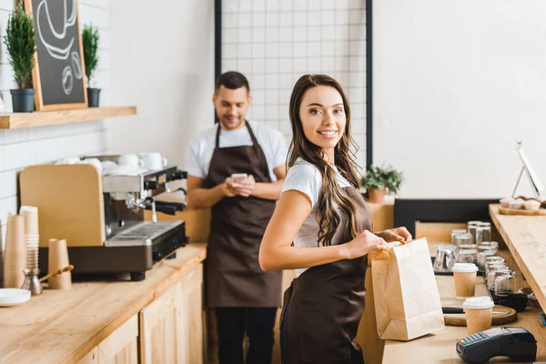 Cajero con bolsa de papel sonriente y barista con teléfono inteligente de pie detrás del mostrador de la barra en la cafetería - foto de stock