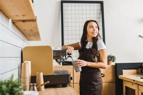 Attraktive brünette Barista hält Tasse und kocht Kaffee mit Maschine im Kaffeehaus — Stockfoto