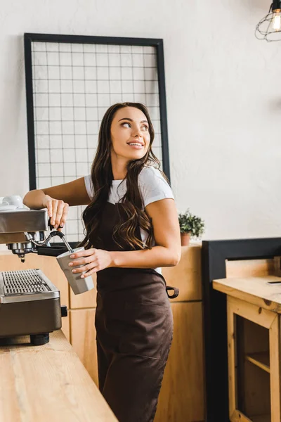 Привлекательная брюнетка бариста в коричневом фартуке держа чашку и кофе с машиной в кофейне — стоковое фото