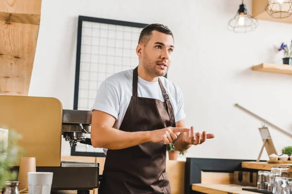 Wütender gutaussehender Barista in brauner Schürze steht neben Kaffeemaschine im Kaffeehaus — Stockfoto