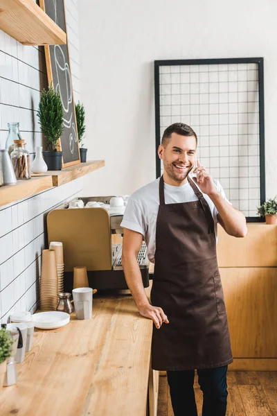 Apuesto cajero en delantal marrón hablando en el teléfono inteligente cerca de la máquina de café y mostrador de bar en la cafetería - foto de stock