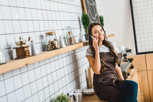 Atractiva cajera en delantal marrón hablando en el teléfono inteligente cerca de la máquina de café y sentado en el mostrador de bar en la cafetería - foto de stock