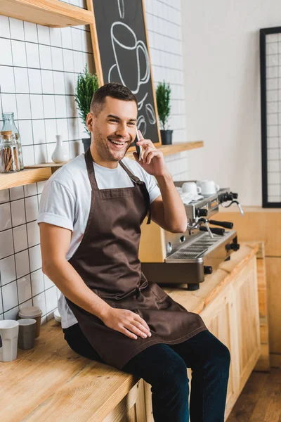 Apuesto cajero en delantal marrón hablando en el teléfono inteligente cerca de la máquina de café y sentado en el mostrador de bar en la cafetería - foto de stock