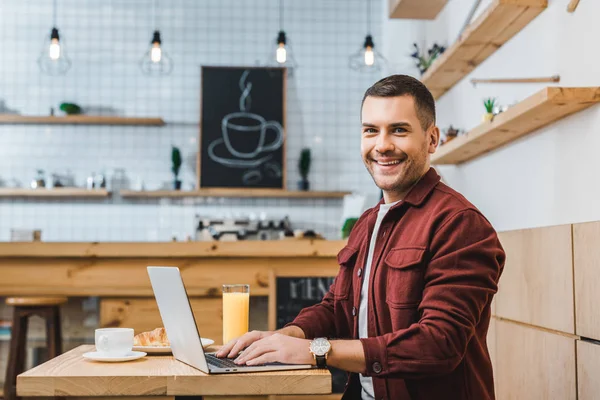 Handsome freelancer sitting at table with laptop and smiling in coffee house — Stock Photo