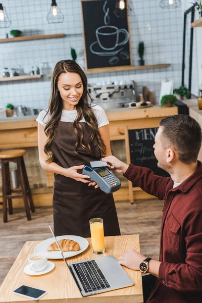 Attractive waitress holding terminal wile handsome man sitting at table with laptop and paying with credit card in coffee house — Stock Photo